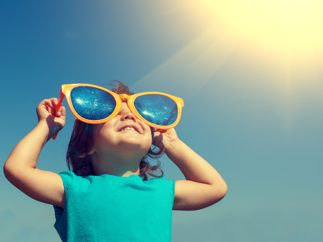 Young child holding large sunglasses over their eyes and looking towards the sun