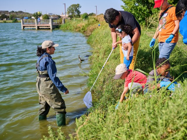 a woman stands in the water in waders while instructing a group of parents and kids during an environmental education lesson at Fish Trap Lake