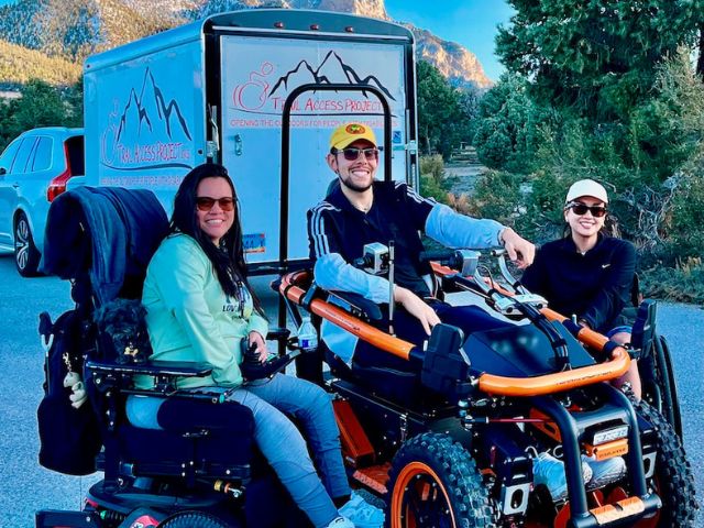 three people in wheelchairs sit smiling in from of a Trail Access Project Trailer