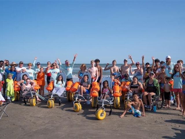 Large group of people of all ages on a beach with some in beach wheelchairs. R.A. Apffel Beach, Park Board of Trustees of the City of Galveston 