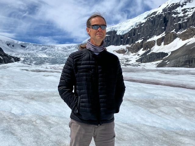 a man stands on a glacier with snop capped mountains in the background