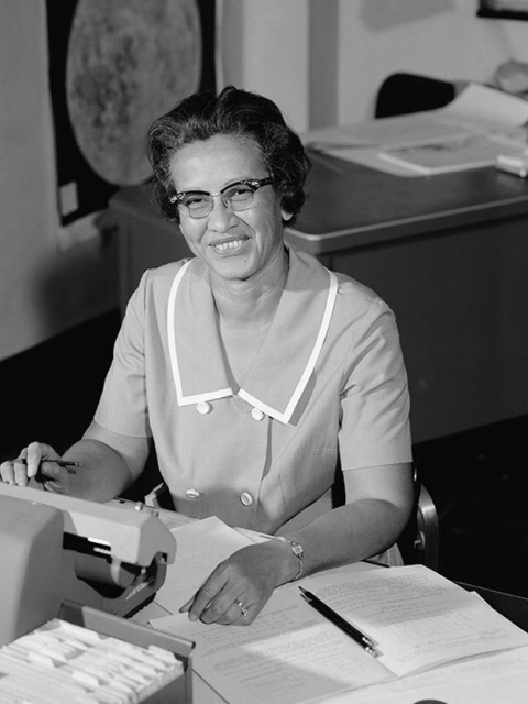 A black and white photo of a woman in glasses sitting at a desk piled high with textbooks and pages of handwritten calculations
