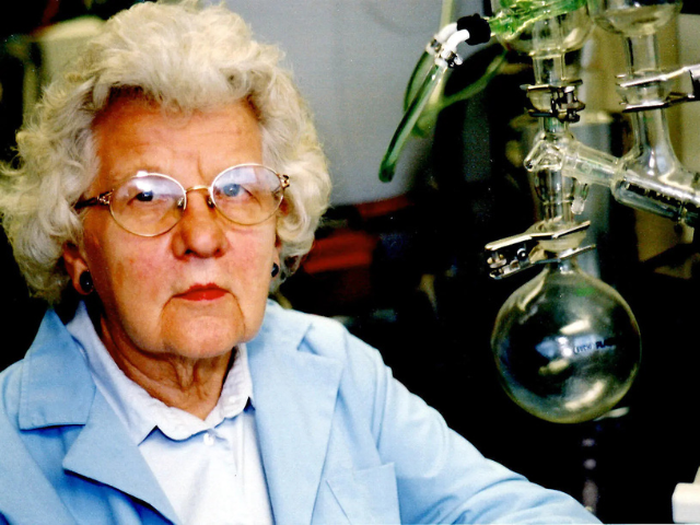 An older woman with gray hair and glasses poses next to a set of chemistry equipment