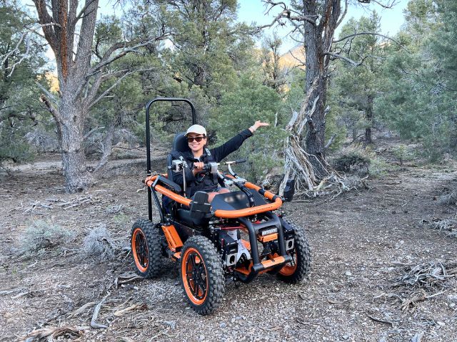 a young woman sits in an all terrain wheelchair on a trail.