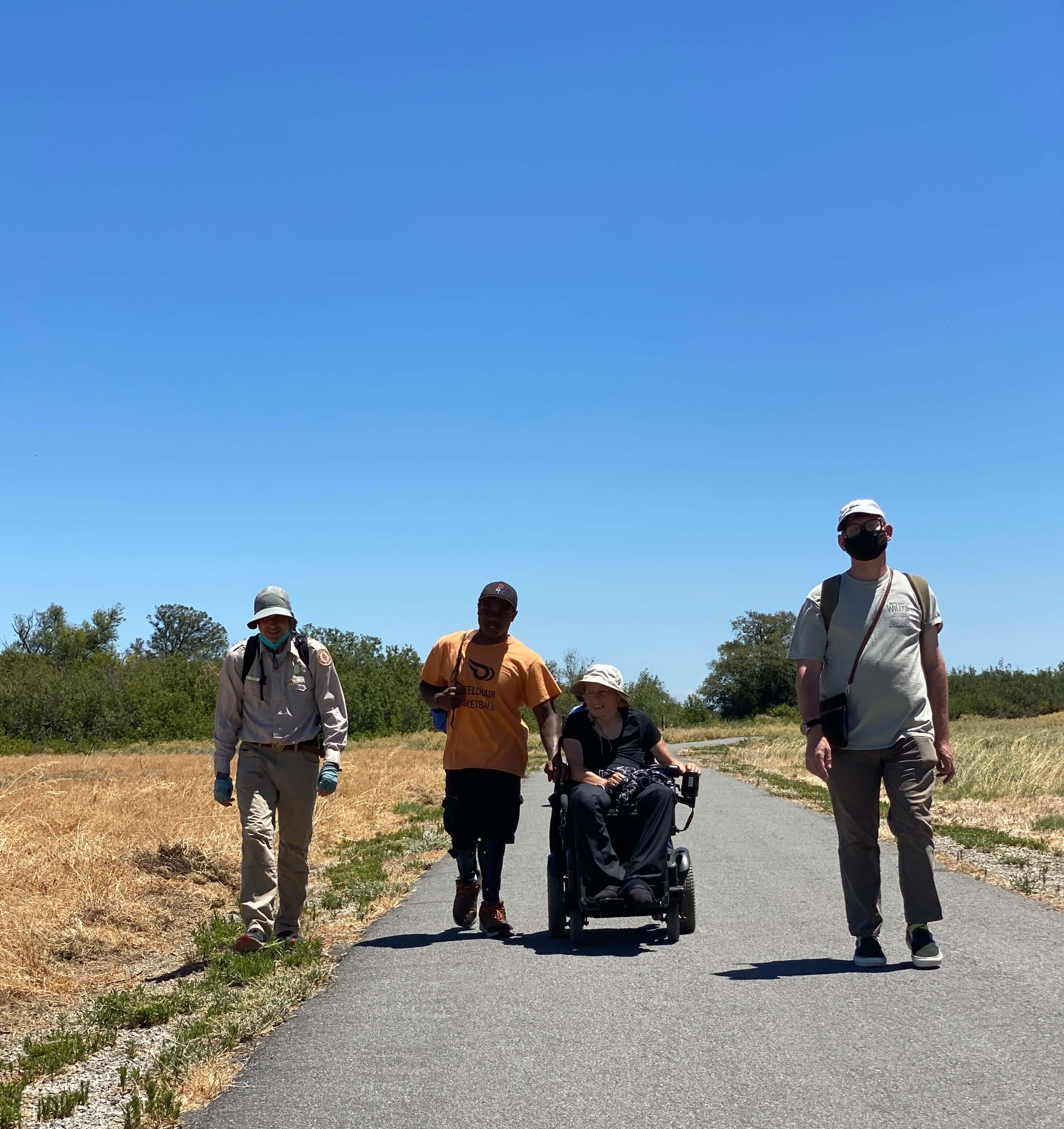 Three individuals, including one in a motorized wheelchair, hike with a park ranger on an asphalt path through dry grasslands at Big Break Regional Shoreline 