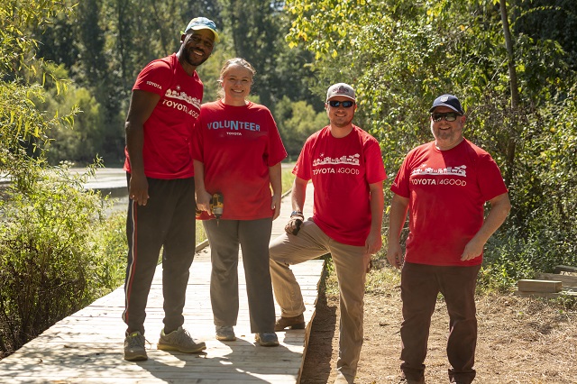 Four smiling people wearing red Toyota shirts, standing outdoors near a wood boardwalk in the woods
