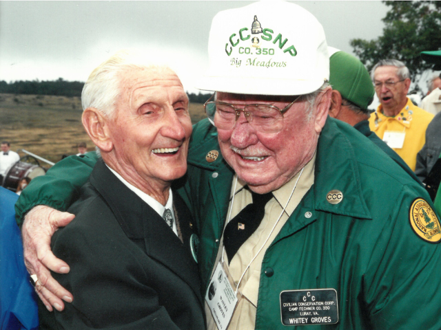 Two men outdoors one with arm around other both smiling