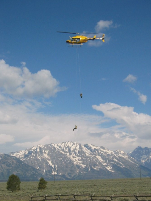 Two people are lowered from a yellow helicopter over a valley with snow-capped mountains in the background.
