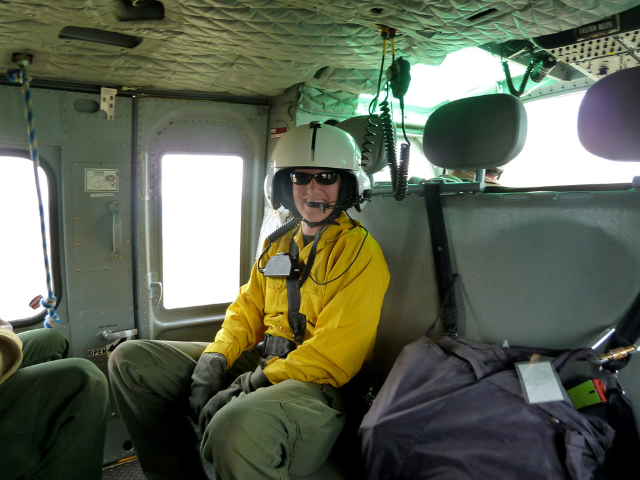 Lorena Williams, wearing a flight helmet and yellow fire shirt, sits inside a helicopter during a fire assignment.