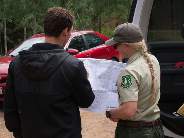A U.S. Forest Service employee reviews a map with another person beside a vehicle in a wooded area.