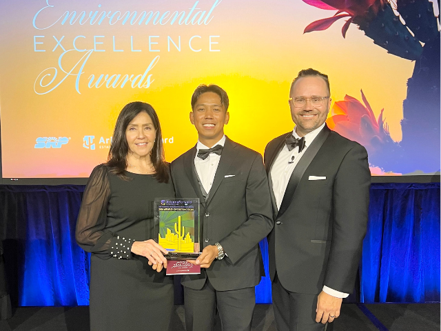 Three people standing in front of a sunset backdrop smiling, one is holding an award.