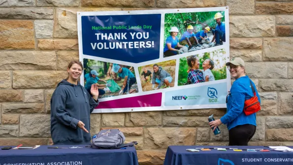 Two national public lands day volunteers stand near a NPLD banner that say Thank You Volunteers