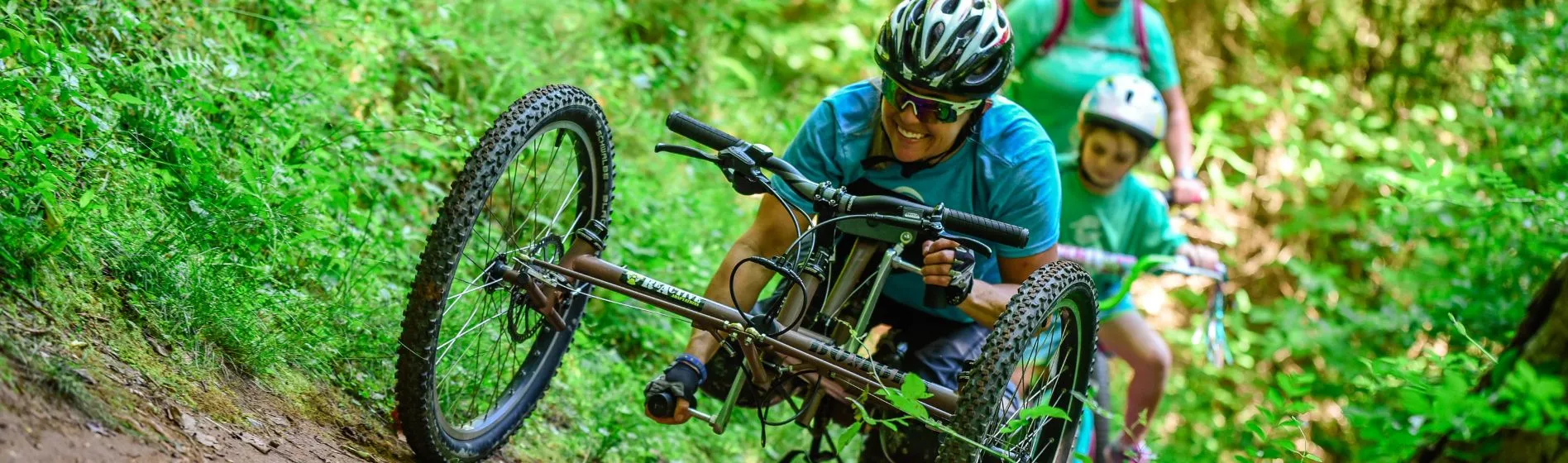 a woman using a wheelchair handcycle moves along a trail in the forest with young girl behind her