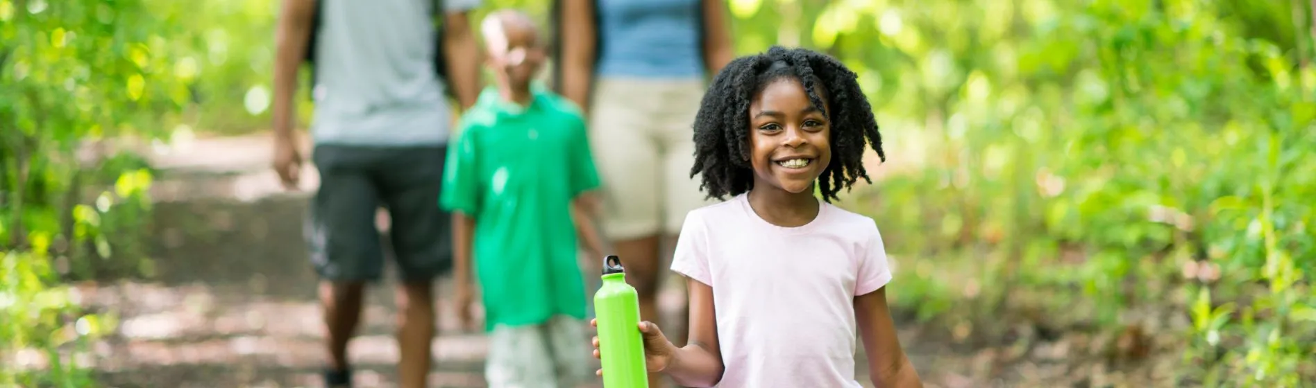 a young girl smiles while walking on a trail in the woods with her family close behind