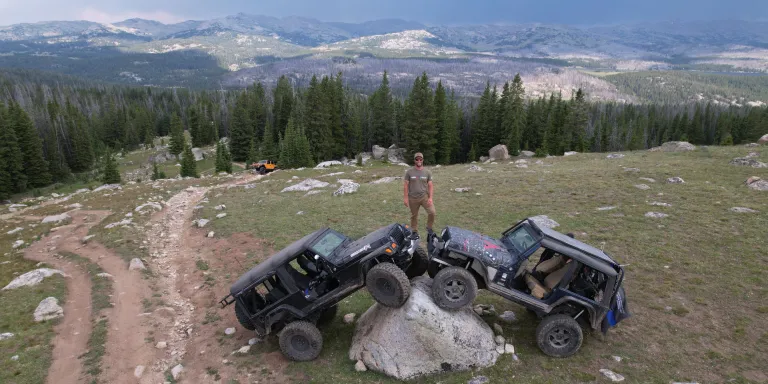 A man stands on top of two off-road vehicles parked on a large boulder. A vast mountain range extends behind him