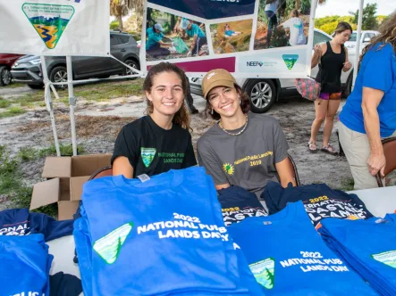 two young women sit at a NPLD event welcome table filled with National Public Lands Day tshrits