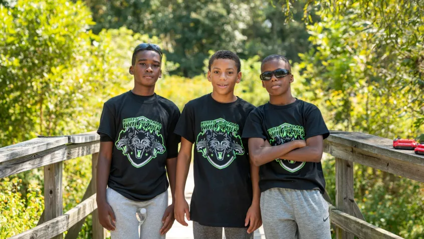 Three young black men stand together on a boardwalk in nature.