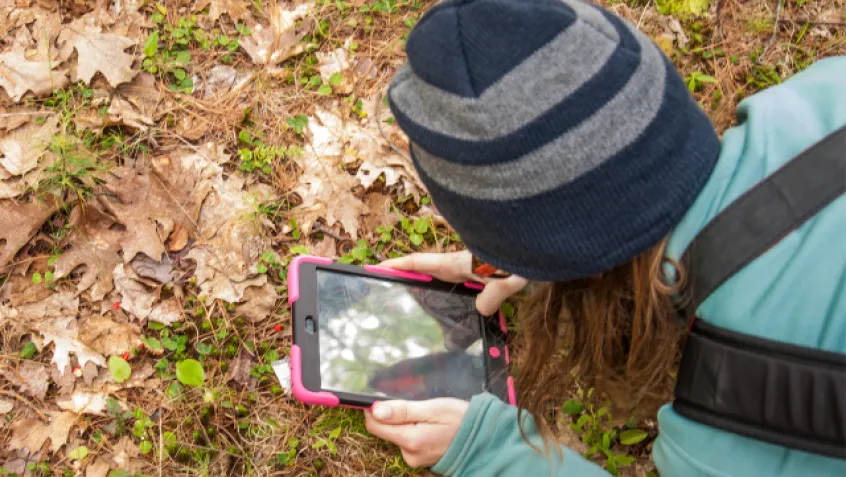 person with long hair wearing hat taking photo with phone of leaves on the ground