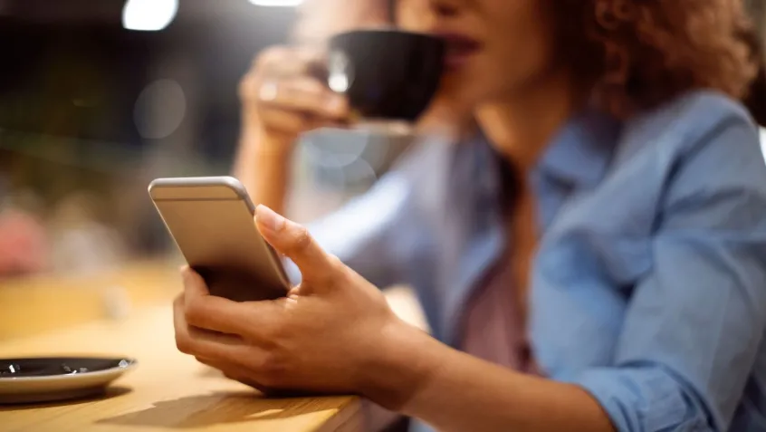 a woman holds a cell phone while reading and drinking from a mug