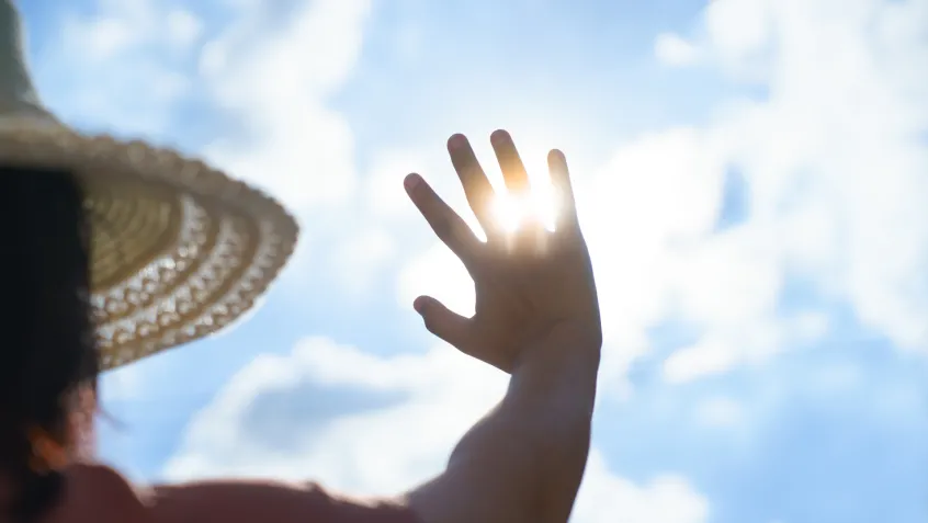 A person wearing a large sun hat holds their hand up to the sky to block the sun's harsh rays.