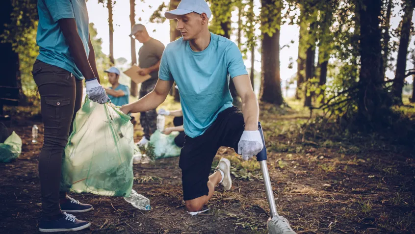 A man with an artificial leg helps pick up trash in a forested setting.