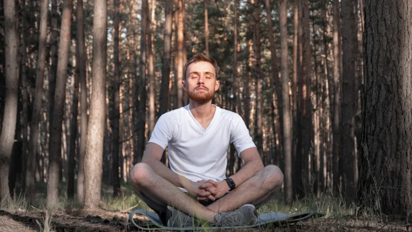 A man sits cross-legged on the ground surrounded by forest