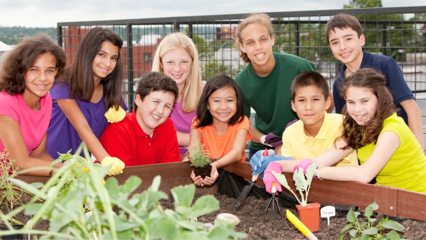 A diverse group of students pose for a photo in front of a raised garden bed in a school garden. A young girl in the center is holding a small plant that is about to be planted in the bed.