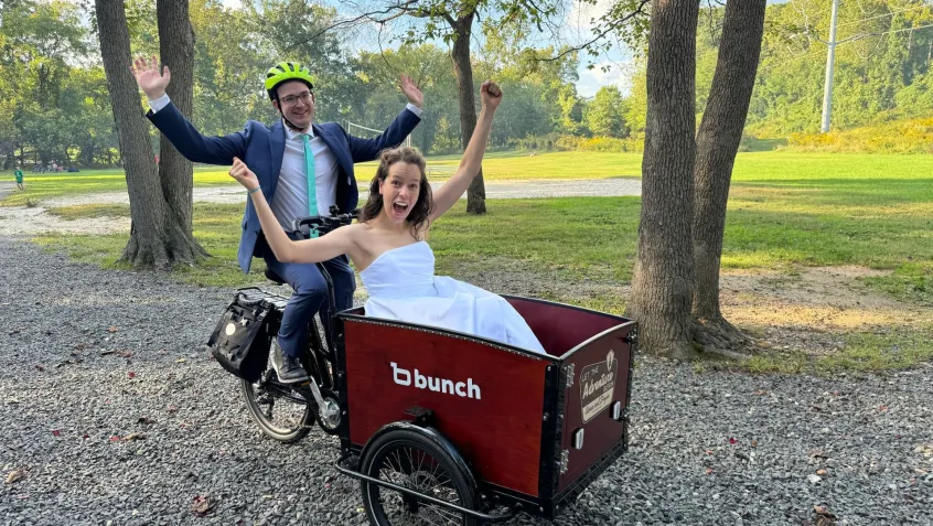 The groom is wearing a bright yellow helmet and sitting on the saddle of a cargo bike. The bridge is in the front basket of the bike. Both of them have their arms raised triumphantly