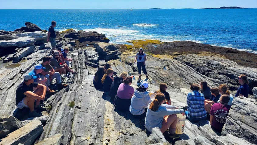 A woman stands on a rocky shoreline speaking to a group of seated college students as they overlook the ocean in the background