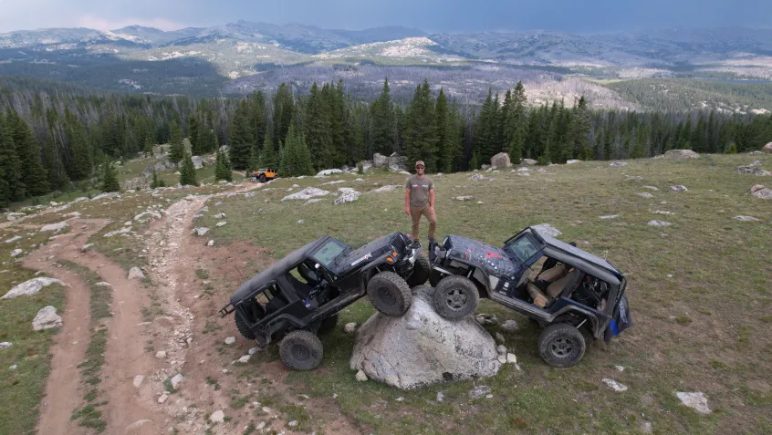 A man stands on top of two off-road vehicles parked on a large boulder. A vast mountain range extends behind him