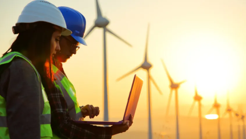 Two people wearing hard hats and reflective vests look at data on a laptop while the sun sets behind a row of wind turbines in the background