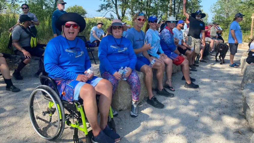 a man using a wheelchair sits with family and friends waiting for a kayak launch