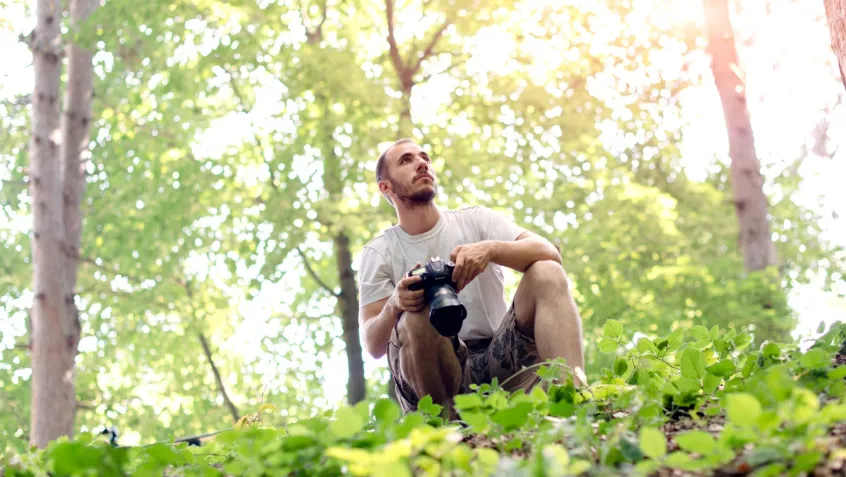 a man with a camera crouches in the forest while looking for birds