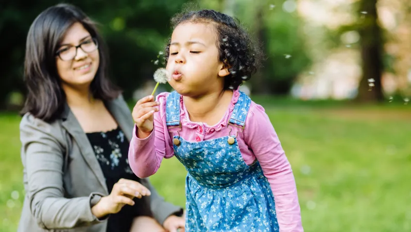 Daughter blowing on a dandelion while her mom watches