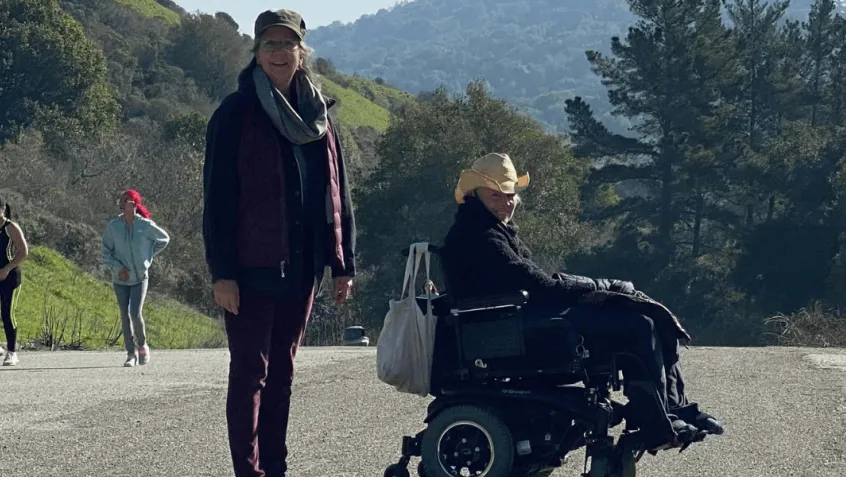 Two woman (one standing, one in a wheelchair) with big smiles hike Wildcat Creek Trail past green hills studded with trees 