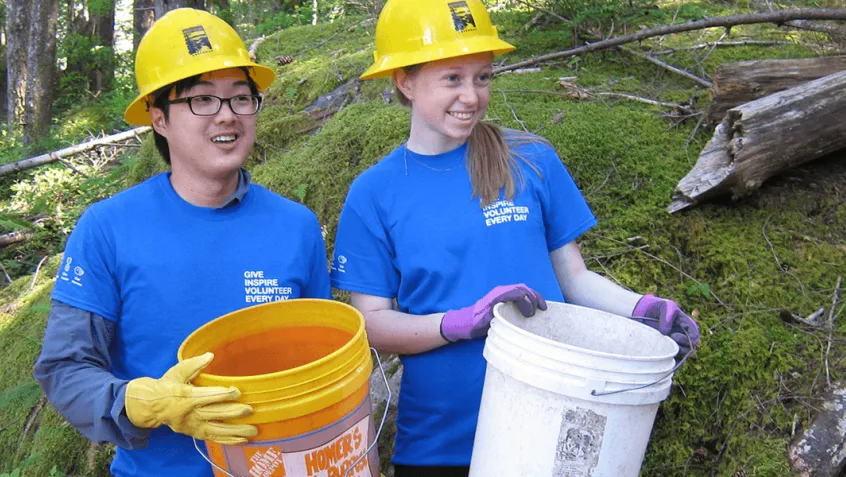 Young woman and man holding buckets and wearing hard hats volunteering outdoors