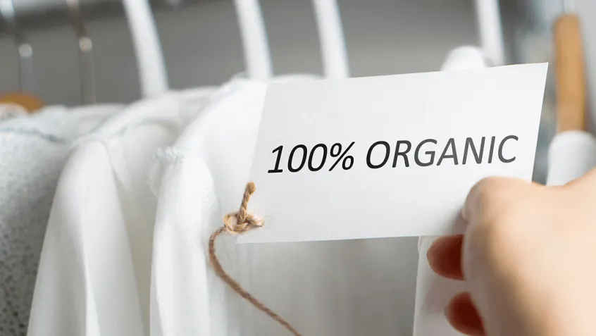 rack of white shirts and hand holding an ecolabel that reads 100% organic