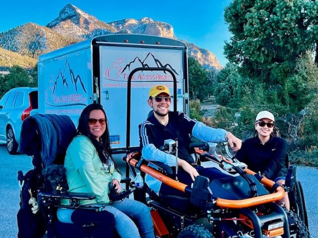 a group of wheelchair users sit smiling with one in an all terrain hopper with mountains in the background.