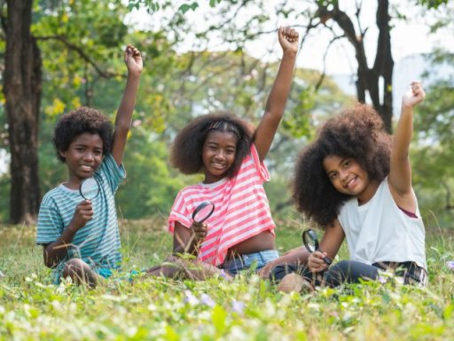 three middle school students with magnifying glass raise their hands while sitting outside for environmental education on public lands