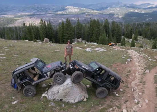 A man stands on top of two off-road vehicles parked on a large boulder. A vast mountain range extends behind him