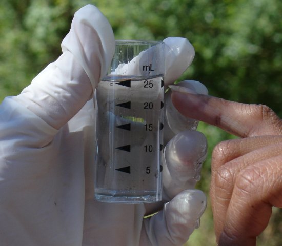 photo of water quality testing, one gloved hand holding a beaker of water and one brown hand point to the beaker.