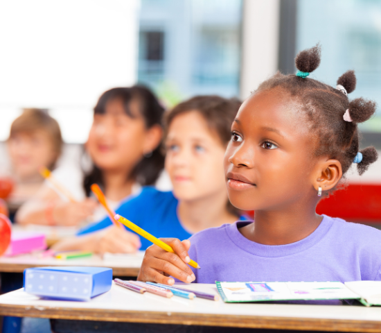 Young girl at desk holding pencil looking beyond frame at teacher