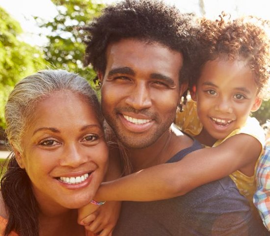 a black man holds his daughter on his back with his wife standing next to him all smiling