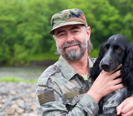 a man in army fatigues holds a dog while outside by a river bank