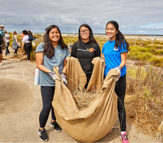 Three young women pose for a photo holding a large bag of invasive plants they've removed from a local habitat.