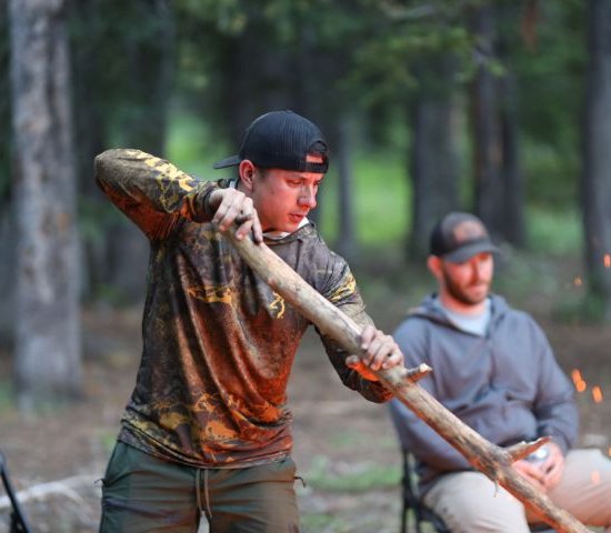 a man wearing camouflage stirs a bonfire while another man sits in a chair behind him in the forest
