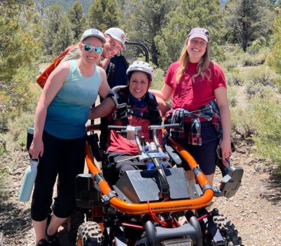 a woman sitting in an all-terrian wheel chair surrounded by her friends on a trail with mountains in the background