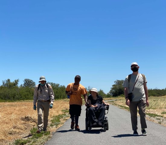 Three individuals, including one in a motorized wheelchair, hike with a park ranger on an asphalt path through dry grasslands at Big Break Regional Shoreline