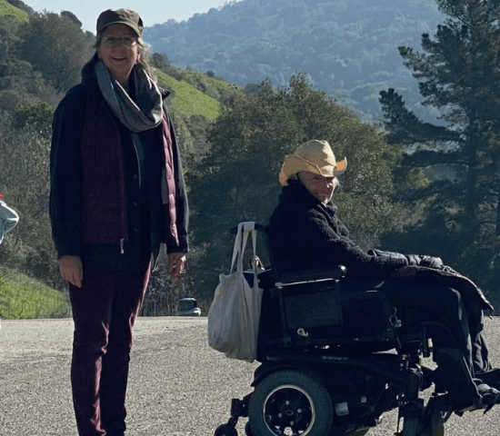 Two woman (one standing, one in a wheelchair) with big smiles hike Wildcat Creek Trail past green hills studded with trees
