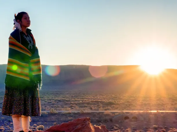Native American young woman wearing traditional dress stands in front of a desert mountain range with sun setting.